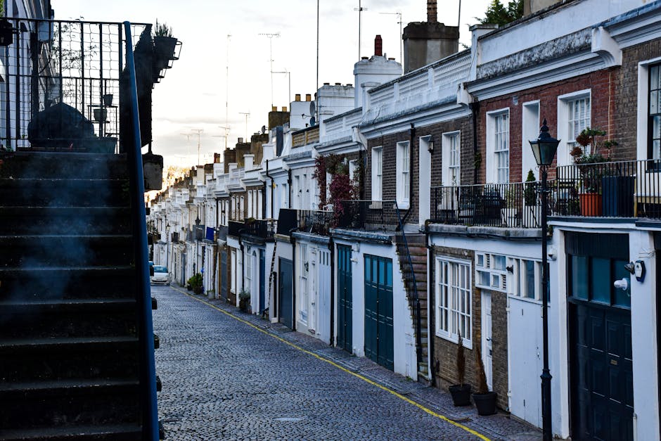 A row of terraced houses in Kentish Town featuring brick and white-painted facades, with front doors and small private balconies adorned with potted plants and outdoor furniture. A narrow cobbled street runs in front of the houses, with a black metal staircase attached to one building on the left side, leading up to an upstairs entrance. The street is lined with black lampposts and small potted plants, with a slight incline visible in the background. Soft natural light from the setting sun illuminates the scene, casting gentle shadows and creating a calm atmosphere, reflecting the typical street environment where home relocation and furniture transport services by companies like Man with Van Kentish Town may take place during local house removals or packing and moving activities.