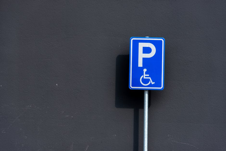 A blue and white parking sign indicating designated disabled parking, mounted on a metal pole against a dark grey exterior wall. The sign features a large white letter 'P' and a wheelchair symbol below it. The sign is situated outdoors in a paved area, with a shadow cast on the wall behind it, suggesting sunlight. The scene depicts a typical urban environment where parking permits, such as Camden Council parking regulations for house removals in NW5, KENTISH TOWN, may apply during a home relocation process. The sign’s placement and visibility are designed to inform drivers about accessible parking restrictions, relevant to moving companies like Man with Van Kentish Town involved in furniture transport and loading activities.