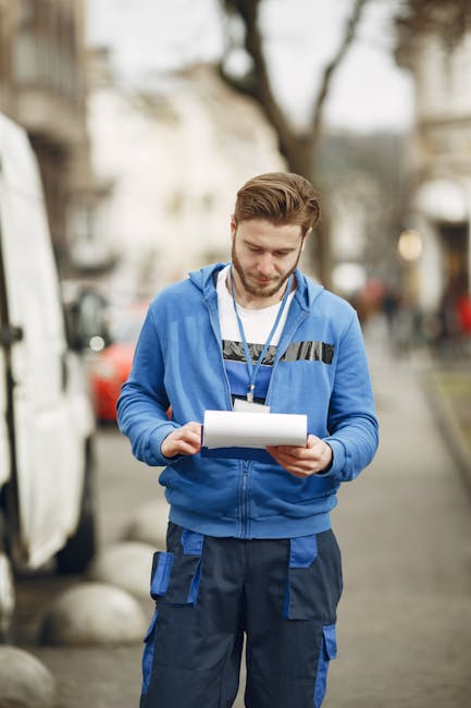 A young man with light brown hair, wearing a blue hoodie with black and grey accents and dark blue work trousers with multiple pockets, stands outdoors on a street during daylight hours. He is holding