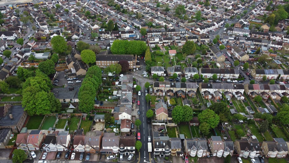 An aerial view of a residential neighbourhood in Kentish Town showing a mixture of terraced houses, semi-detached homes, and detached properties, all surrounded by lush green trees and small gardens. Cars are parked along the narrow streets, and the rooftops display a variety of materials, including slate and tile. The image captures the quiet street scene during daytime, with the houses neatly aligned and the foliage providing a vibrant contrast. This setting illustrates the typical environment for house removals and home relocation processes, where furniture transport and packing activities take place within a suburban area. The photograph emphasizes the compact urban layout, ideal for professional removals services like those offered by Man with Van Kentish Town, assisting families with efficient and careful moving logistics within this community.