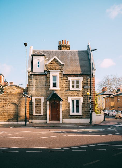 A multi-storey brick house with a gabled roof and several chimney stacks, situated on a paved street in a residential area. The building has white-framed windows and a brown wooden front door beneath a small porch canopy. To the left of the house, there is a brick wall with an arched opening, and to the right, there are parked cars and other residential buildings. The sky is blue with some clouds, and a streetlamp is positioned on the pavement in front of the property. This scene represents a typical home exterior in an urban neighbourhood, relevant to house removals and moving services such as those offered by Man with Van Kentish Town, emphasizing the typical residential environment for furniture transport and home relocation processes.