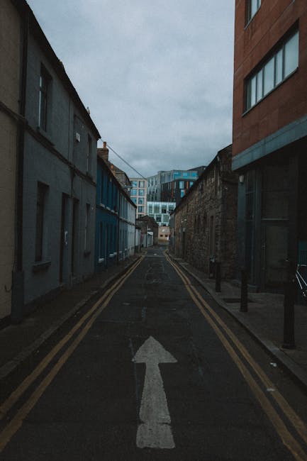 A narrow urban street in Kentish Town with a dark, cloudy sky overhead. On the left, there are small, weathered terraced buildings painted in shades of blue and grey, some with visible windows and doors. On the right, a modern building with large glass windows and a brick façade is visible. The street is marked with a white arrow pointing straight ahead, and double yellow lines run along both sides of the asphalt pavement. Several black bollards are positioned along the pavement edge on the right. The scene suggests a location suitable for house removals or furniture transport, with no vehicles or people present, emphasizing a typical street where local moves or loading for a home relocation might take place. This setting aligns with the services offered by Man with Van Kentish Town, providing contextual visual support for house removals on narrow streets in NW5.