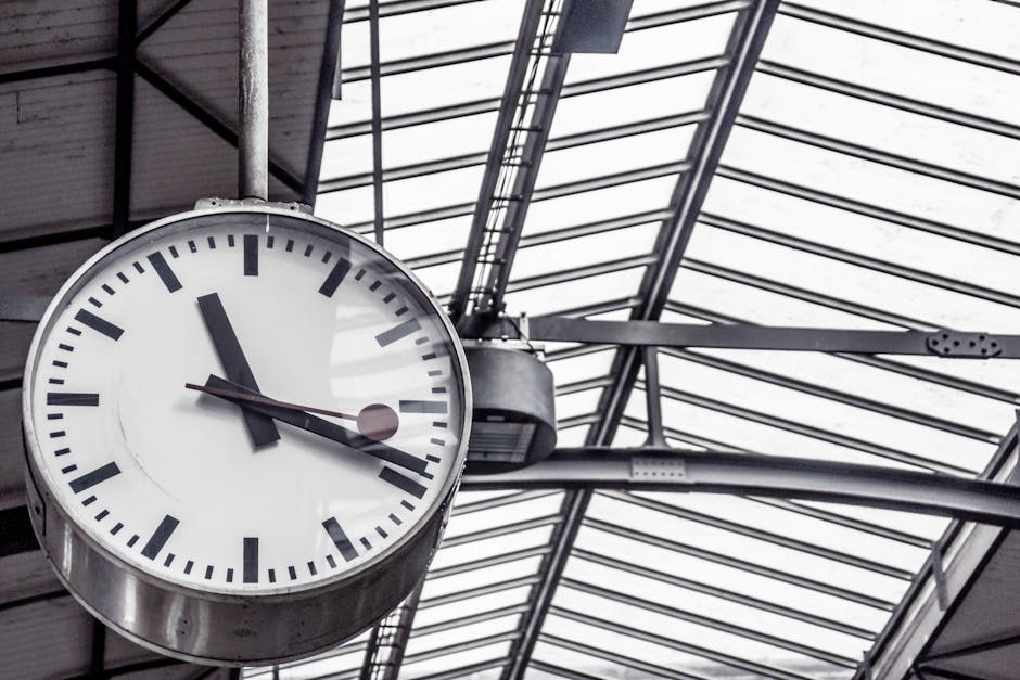 A large round analog clock with a white face and black hour and minute hands, mounted on a ceiling at Kentish Town station. The clock shows approximately 4:55. Above the clock, there is a glass and metal roof structure with a grid pattern, allowing natural light to illuminate the area. The surrounding environment features metal beams and support structures, typical of a railway station. The image captures the interior space where station staff, such as those from Man with Van Kentish Town, may organize or coordinate home relocation and furniture transport services, potentially involving loading or unloading processes for house removals, within a well-lit station environment.