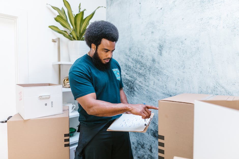 A professional mover from Man with Van Kentish Town wearing a navy blue T-shirt and black apron, holding a clipboard with notes, appears to be inspecting or preparing for a house relocation. He is sta