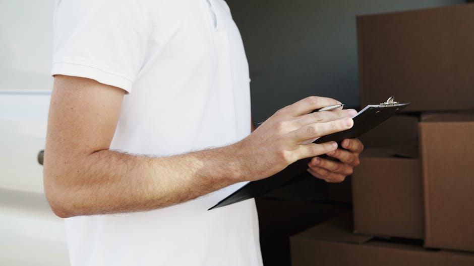 A professional mover from Man with Van Kentish Town wearing a navy blue T-shirt and black apron, holding a clipboard with notes, appears to be inspecting or preparing for a house relocation. He is sta