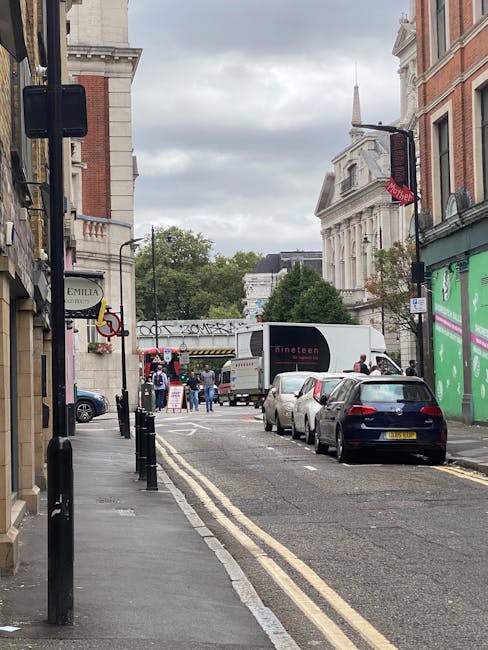 A narrow street in Kentish Town with parked cars along the curb and several pedestrians walking towards a building with a white facade and columns. A white box truck with the logo 'Dinnet' is positioned in the middle of the road, likely involved in a home relocation or furniture transport process. The street is lined with buildings featuring brick and stone exteriors, and there are shop signs visible on the left side. Double yellow lines run along the edge of the road, and black bollards are set on the pavement to prevent vehicle encroachment. Overhead, the sky is overcast with grey clouds, and streetlights and traffic signals are mounted on metal poles. The environment suggests a typical urban setting suitable for professional removals and moving services, as supported by the context of [PAGE_TITLE] and the presence of a moving vehicle prepared for loading or unloading furniture.