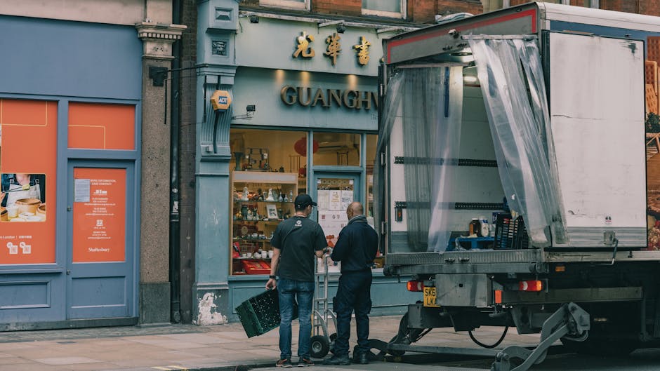 Two men are engaged in a home relocation activity outside a shop with a sign that reads 'Guangh' above the entrance. One man, dressed in dark clothing, stands facing the other man who is wearing a black cap and navy jacket; they are discussing the moving process. A large moving truck with an open side panel is parked on the pavement nearby, with the side panel covered by a transparent plastic sheet for protection during loading. Inside the truck, several black plastic crates and items are visible, and the truck is positioned close to the shop entrance for loading or unloading household belongings. The scene also shows cardboard boxes and packing materials, indicating packing and moving operations, possibly as part of an emergency or same-day removal service. The pavement and building facade are typical of an urban street environment in Kentish Town, and the overall lighting suggests daytime conditions. This image relates to professional removals and furniture transport services offered by Man with Van Kentish Town, supporting efficient and organized house removals.