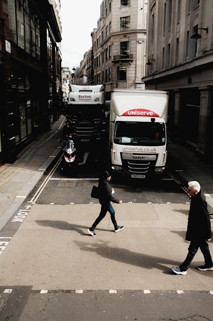 A narrow street in Kentish Town with parked cars along the curb and several pedestrians walking towards a building with a white facade and columns. A white box truck with the logo 'Dinnet' is positioned in the middle of the road, likely involved in a home relocation or furniture transport process. The street is lined with buildings featuring brick and stone exteriors, and there are shop signs visible on the left side. Double yellow lines run along the edge of the road, and black bollards are set on the pavement to prevent vehicle encroachment. Overhead, the sky is overcast with grey clouds, and streetlights and traffic signals are mounted on metal poles. The environment suggests a typical urban setting suitable for professional removals and moving services, as supported by the context of [PAGE_TITLE] and the presence of a moving vehicle prepared for loading or unloading furniture.