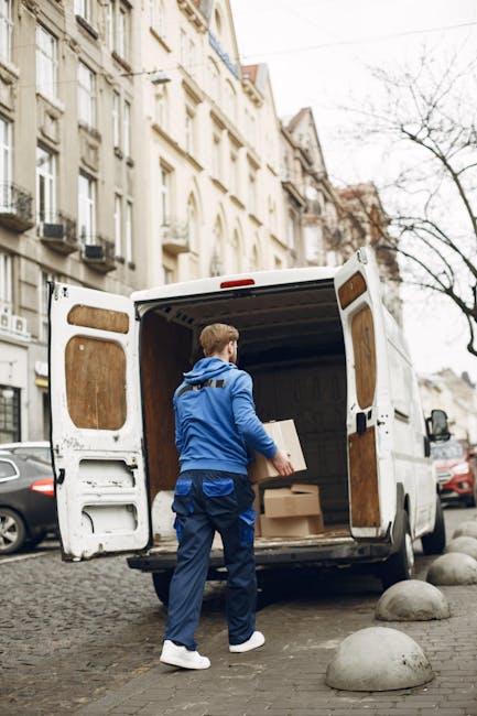 A man wearing a blue jacket, blue trousers, and white sneakers is loading cardboard boxes into the open back of a white panel van parked on a cobblestone street in Kentish Town. The van’s interior walls are lined with wooden panels, and several packed boxes are visible inside. The surrounding environment features multi-storey residential buildings with classic architectural facades, some with balconies and black window frames, and a few parked cars along the street. A leafless tree stands nearby, indicating a colder season. The scene depicts a home relocation or furniture transport process, with the man preparing items for a move as part of a professional removal service provided by Man with Van Kentish Town, suitable for addressing relocation needs when lifts are unavailable.