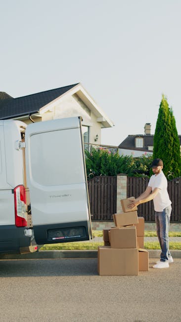 A man wearing a white t-shirt and jeans is loading cardboard boxes into the open rear door of a white van parked on a residential street. The boxes are of various sizes and appear to be securely packed, some stacked on the pavement beside him. The background features a suburban house with a gabled roof, a garden with green bushes and a tall, narrow evergreen tree, and a wooden fence. The scene is well-lit with natural daylight, and the image captures the process of home relocation or furniture transport, consistent with professional removals services provided by Man with Van Kentish Town.