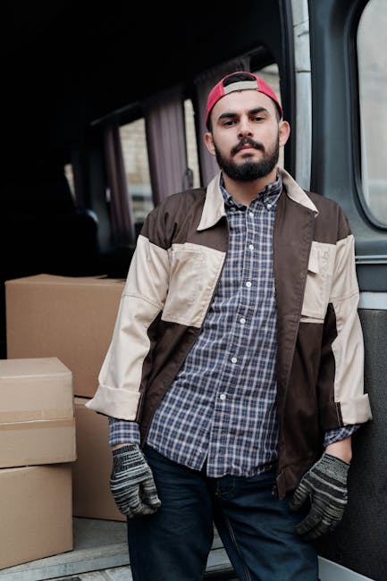 A man wearing a blue jacket, blue trousers, and white sneakers is loading cardboard boxes into the open back of a white panel van parked on a cobblestone street in Kentish Town. The van’s interior walls are lined with wooden panels, and several packed boxes are visible inside. The surrounding environment features multi-storey residential buildings with classic architectural facades, some with balconies and black window frames, and a few parked cars along the street. A leafless tree stands nearby, indicating a colder season. The scene depicts a home relocation or furniture transport process, with the man preparing items for a move as part of a professional removal service provided by Man with Van Kentish Town, suitable for addressing relocation needs when lifts are unavailable.